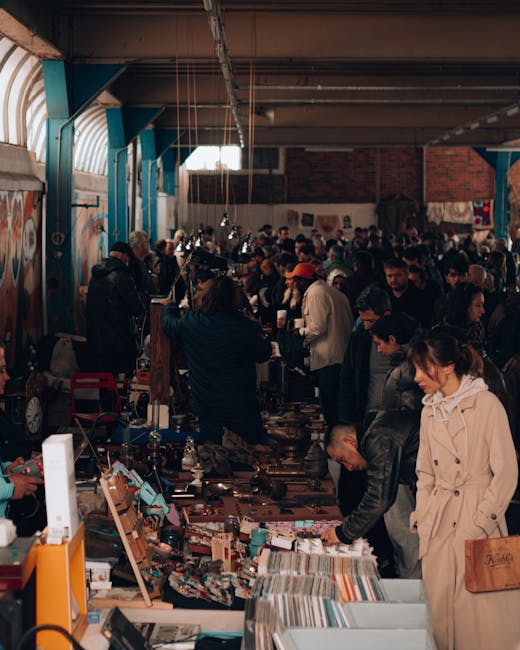 A busy outdoor market scene beneath a large white canopy with multiple stalls displaying an array of secondhand goods including wooden furniture, lamps, and miscellaneous household items. Several vend