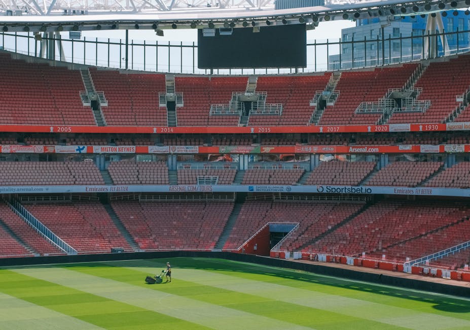 An empty stadium seating area with rows of weathered, orange plastic seats arranged in slight diagonal alignment, some showing signs of wear such as scratches and faded patches. The seats are mounted