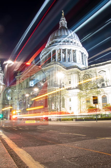 A wide street scene in central London featuring historic white and grey stone buildings with ornate architectural details lining both sides, and St. Paul's Cathedral prominently visible in the backgro